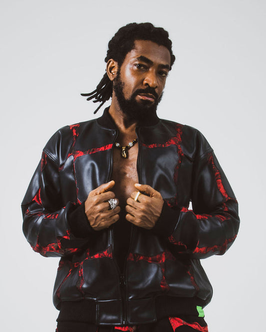 A man modeling a black leather bomber jacket and matching black pants with a red adire pattern, posed against a light gray studio background.