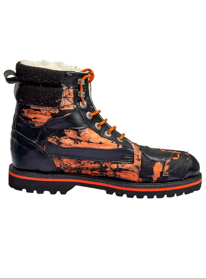 A high-top lace-up boot with a glossy black and orange distressed paint-splatter pattern, featuring orange laces, a black heel pull tab, and a thick black lug sole with a bright orange stripe.