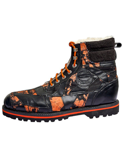 A high-top winter boot with a black and orange splatter patterned leather upper, orange laces, a black sole with a bright orange stripe, and a white faux-fur lining visible at the collar, shown against a plain white background.