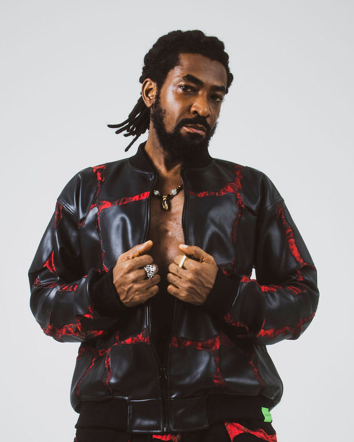 A man modeling a black leather bomber jacket and matching black pants with a red adire pattern, posed against a light gray studio background.