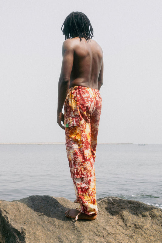Topless man standing on a rock looking out to sea, wearing orange tie-dye combat pants