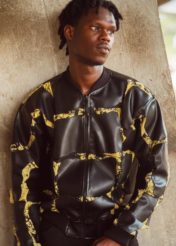 A man wearing a black leather bomber jacket with a distressed yellow pattern, leaning against a textured concrete pillar.