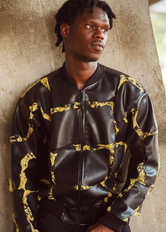 A man wearing a black leather bomber jacket with a distressed yellow pattern, leaning against a textured concrete pillar.