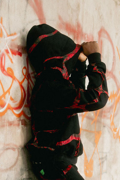 Person in black hoodie and pants leaning against a graffiti-covered wall with a skateboard.