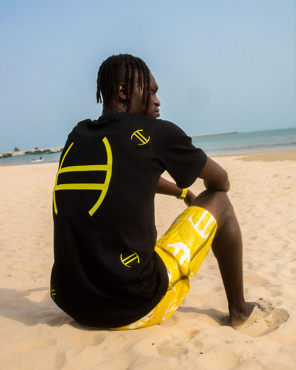 Person sitting on a beach wearing a black t-shirt with yellow designs and yellow shorts.